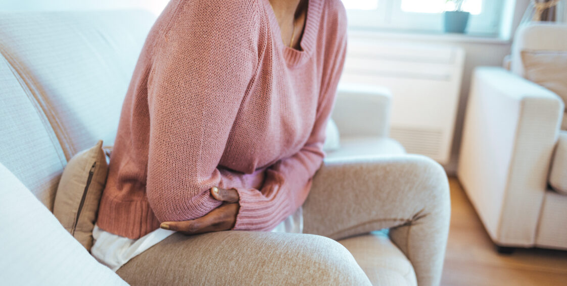 A woman, wearing a pink sweater and beige pants, sits on a couch. She is holding her stomach in pain.